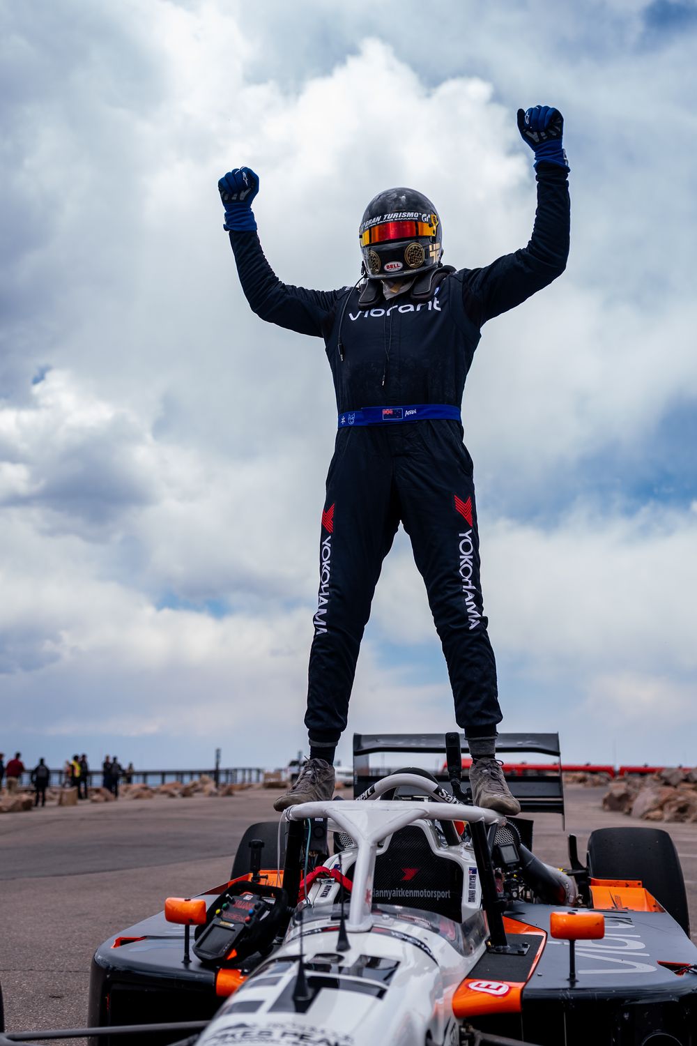 Racing driver in black suit stands triumphantly on top of a race car with arms raised, celebrating a victory under cloudy skies.