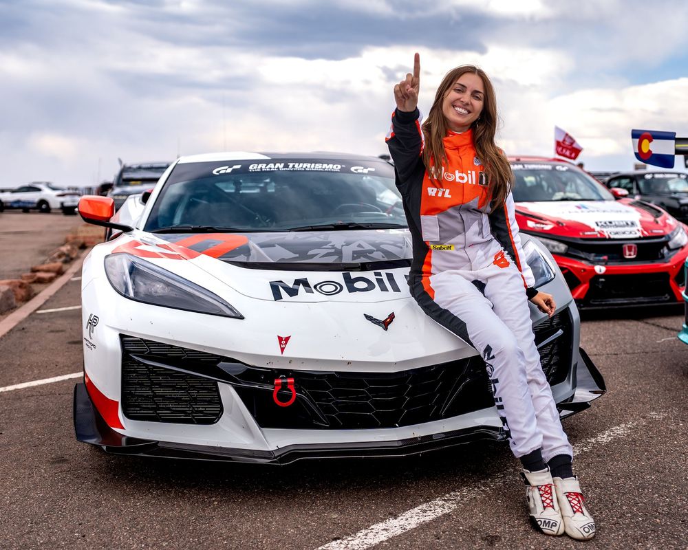 Female racing driver poses with number one gesture beside a Mobil-sponsored Corvette race car at a motorsport event.
