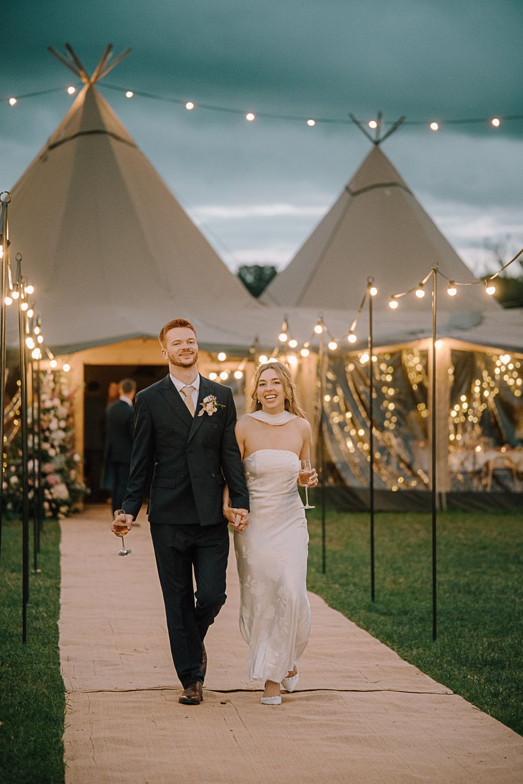 Bride and groom walk hand-in-hand along a lit pathway with festoon lights and tipi tents at dusk.