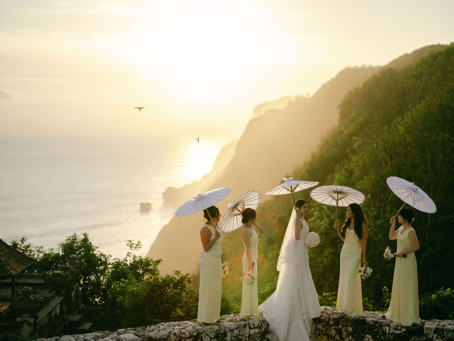 Bridesmaids in white dresses holding parasols stand on a Bali cliffside overlooking the ocean at sunset.