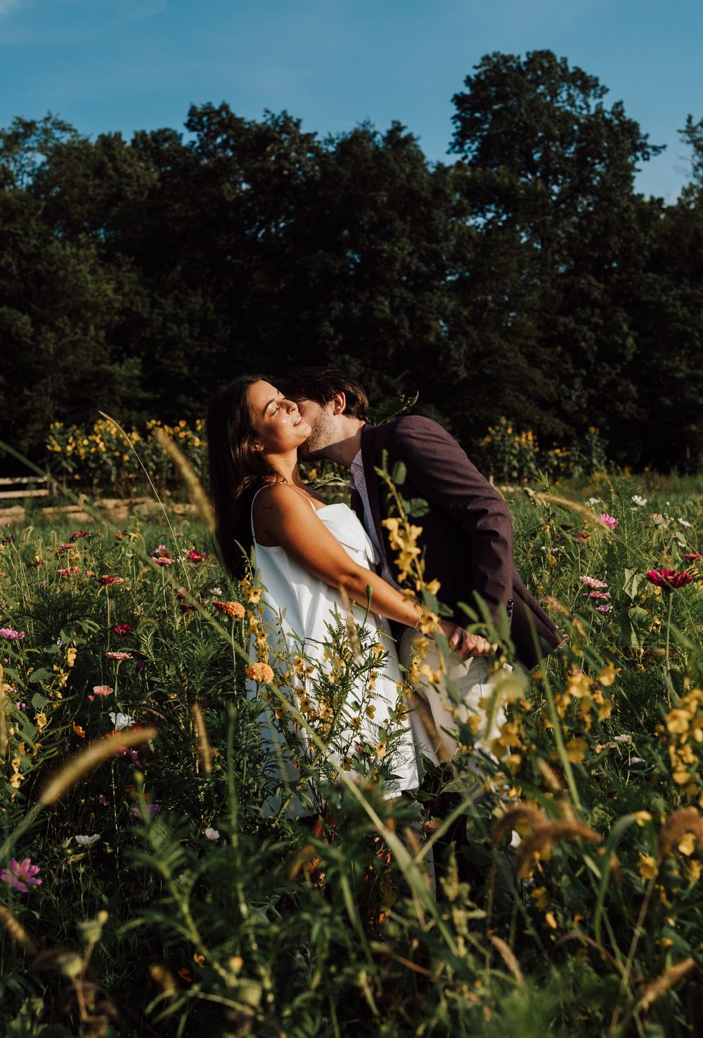 Couple kissing in a colorful wildflower field at golden hour with lush green trees in the background.