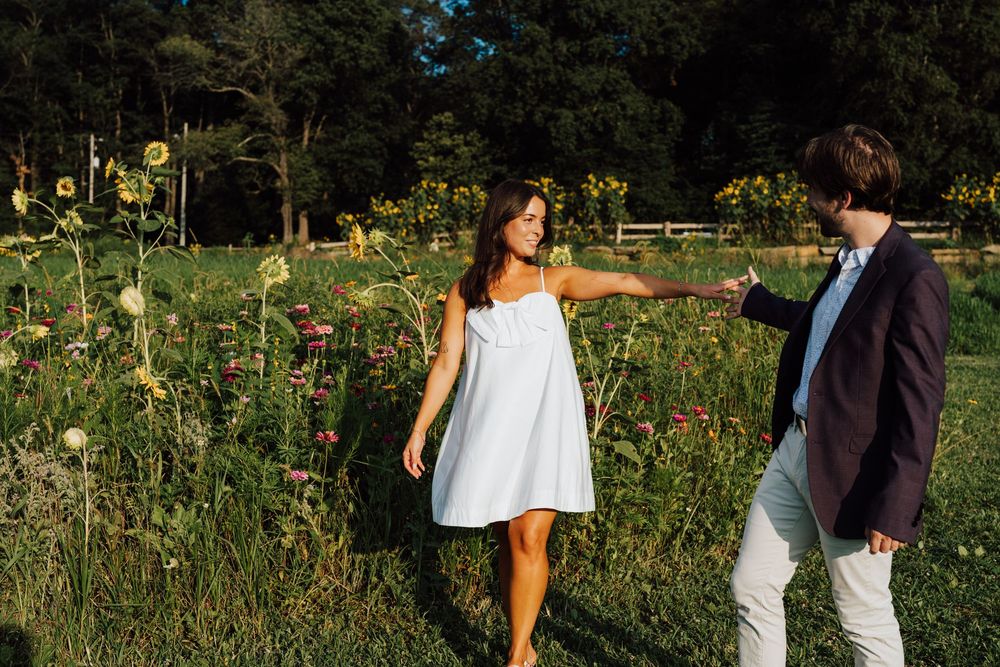 Engaged Couple walking through Wildflower Overlook wildflower meadow at golden hour, woman in white dress reaching back to hold mans hand.