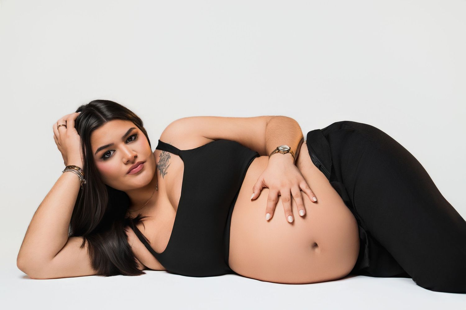 Pregnant woman in black outfit lying down, cradling baby bump during a maternity photoshoot on white background.