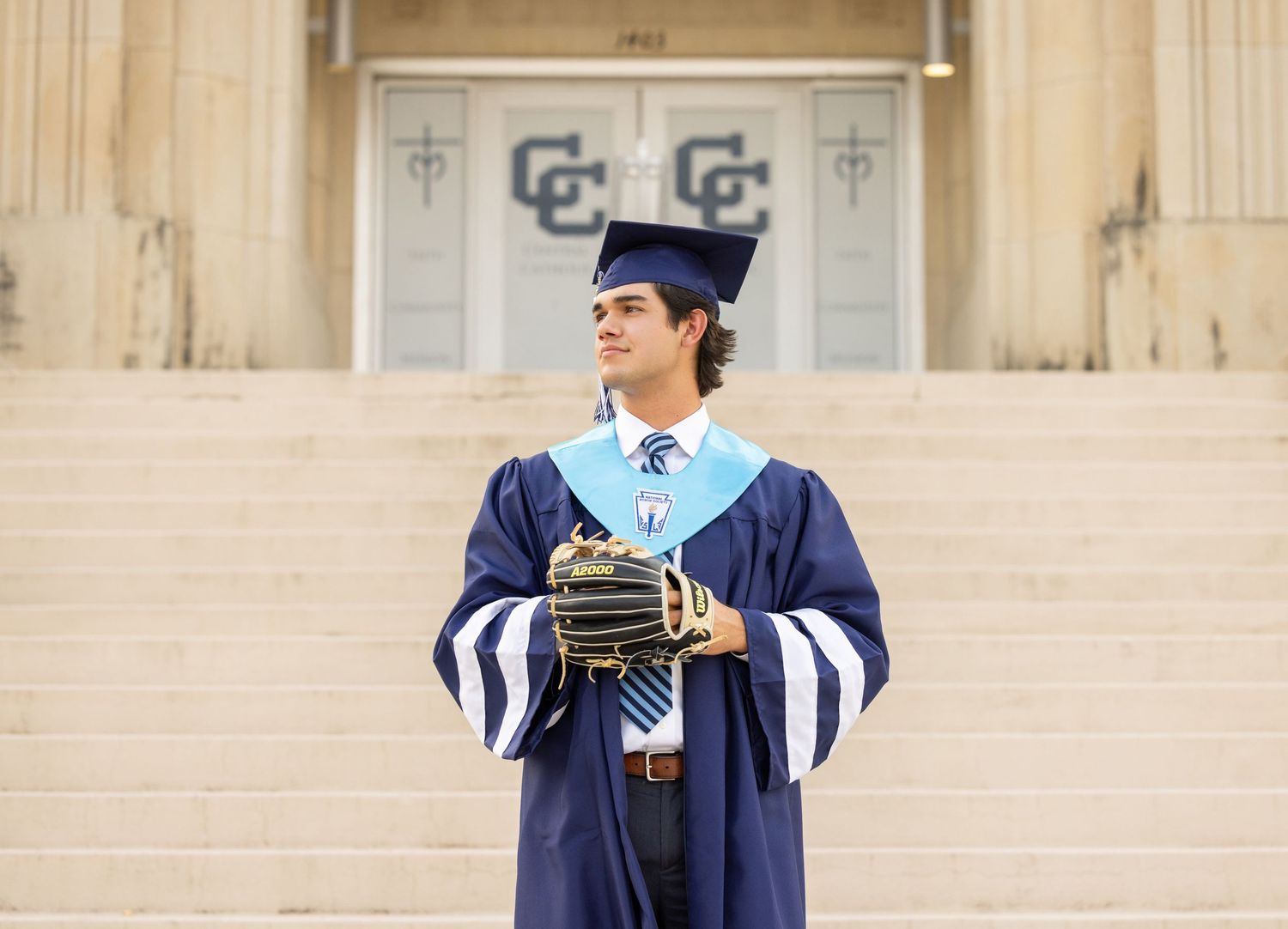 High school graduate in navy blue cap and gown holds baseball glove on steps of school building.