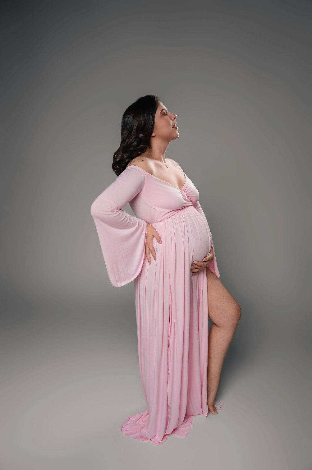Pregnant woman in pink off-shoulder maternity gown posing outdoors on a garden path with lush hedges.