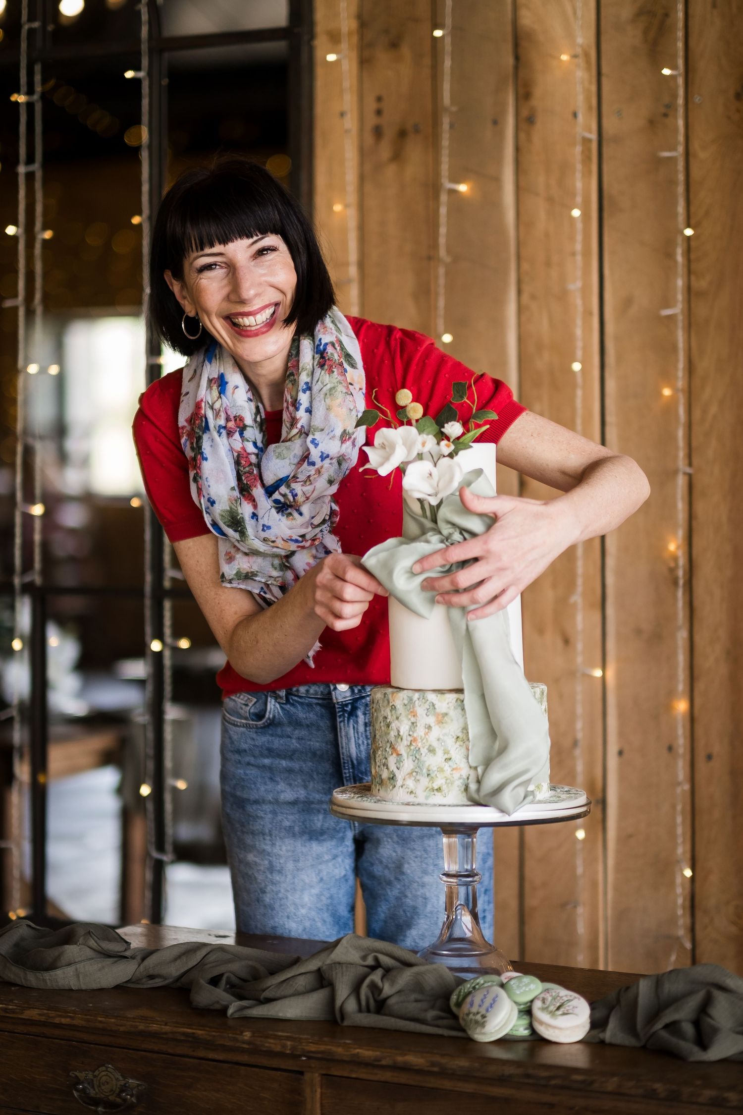 Smiling woman in red top decorating elegant white wedding cake with flowers on wooden table.