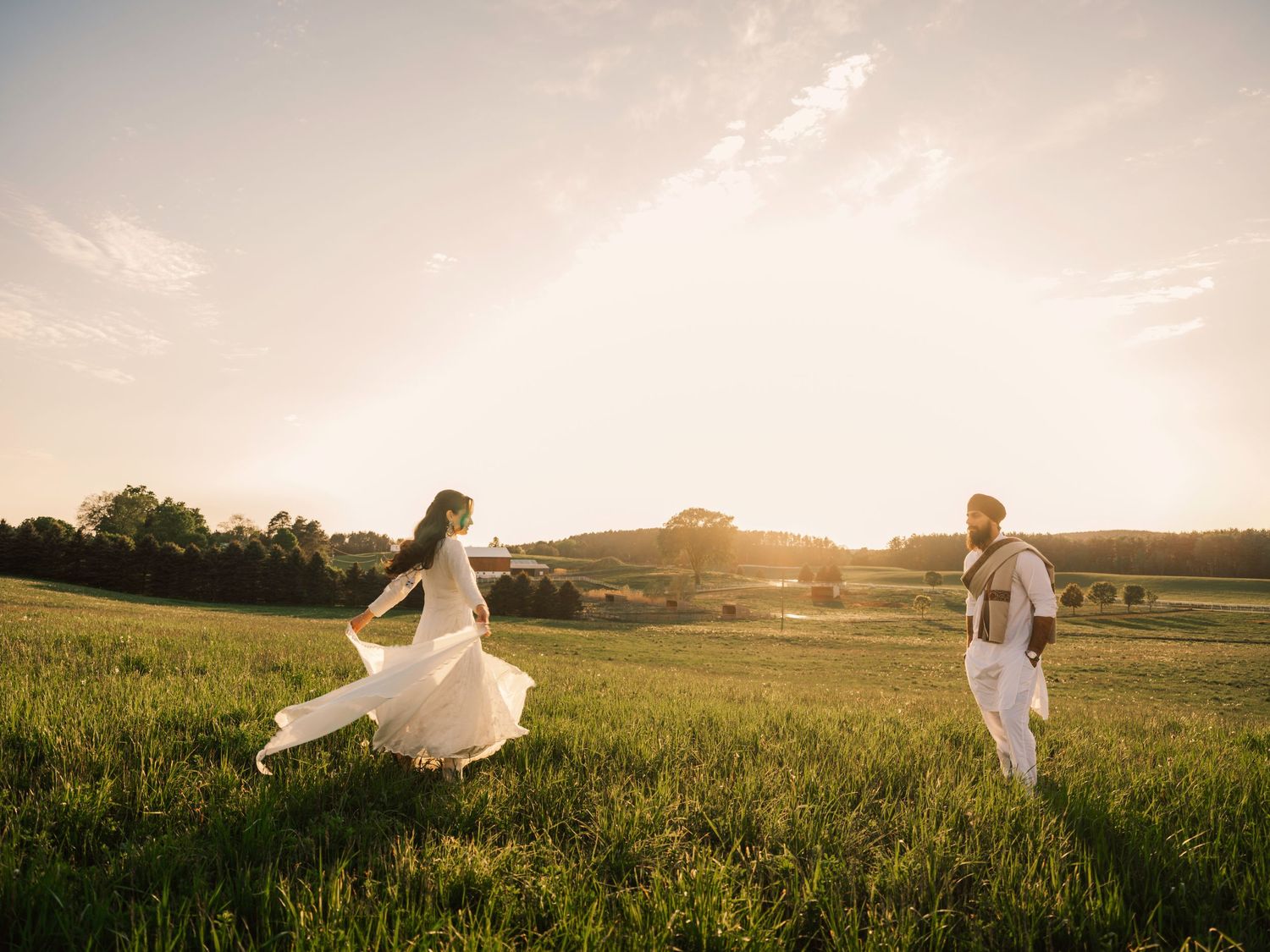 Couple in white outfits running through a lush green field at golden sunset hour.