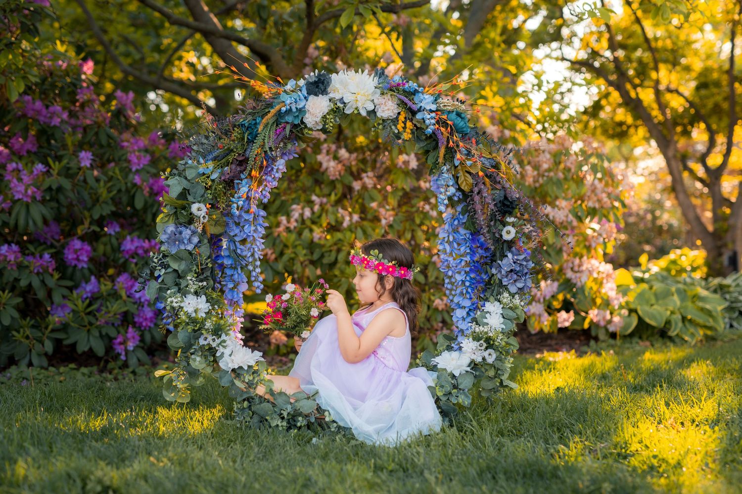 Brown-haired girl wearing a flower crown and holding a bouquet in a garden-inspired portrait.