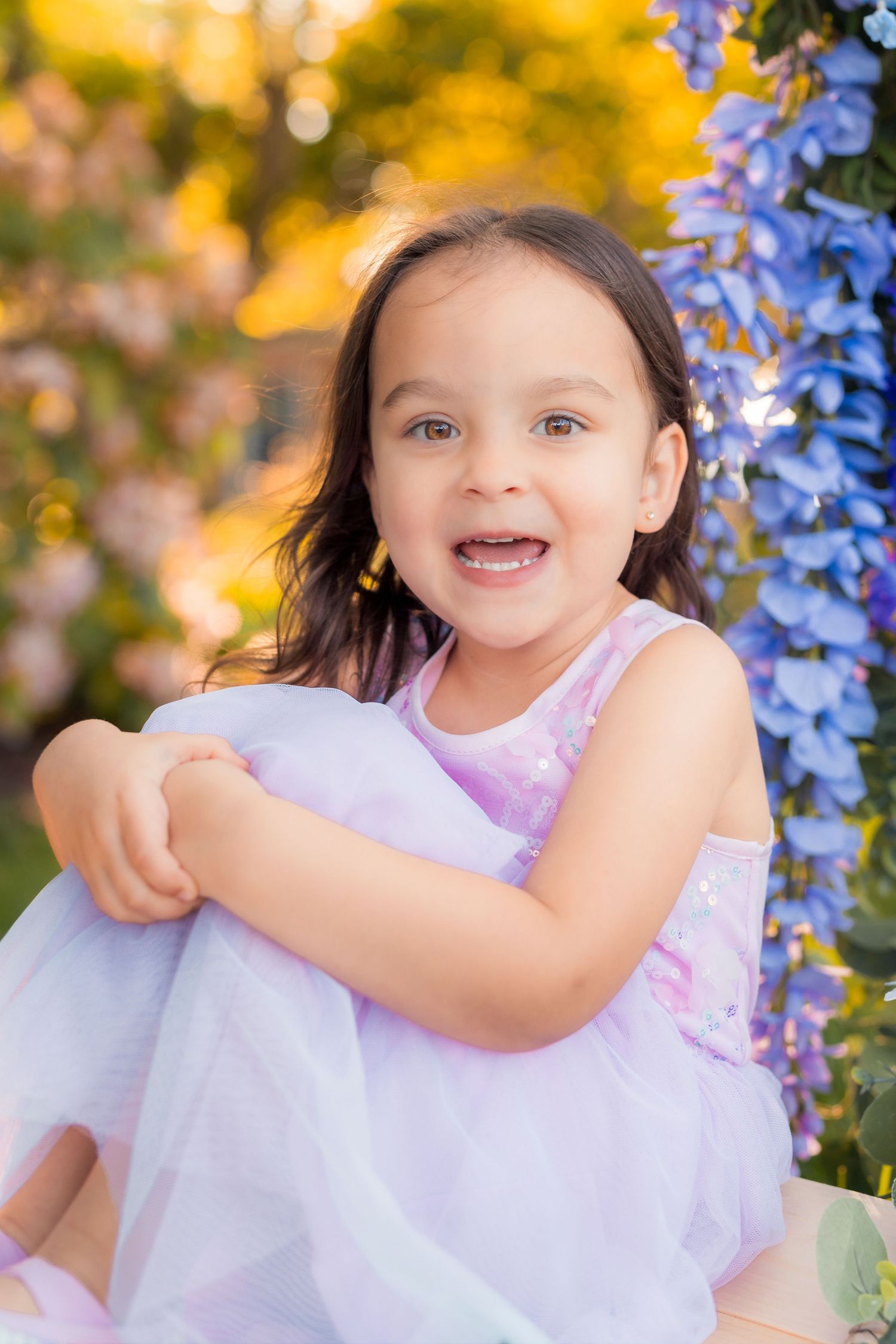 Close-up of a smiling brown-haired girl surrounded by colorful spring flowers.