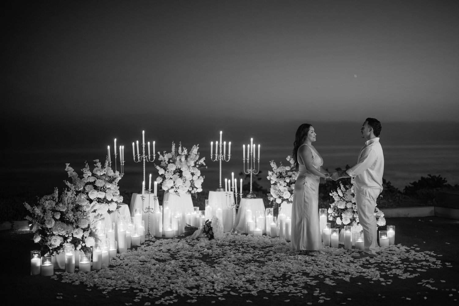 Romantic black and white photo of a couple exchanging vows surrounded by candles and floral arrangements at dusk.