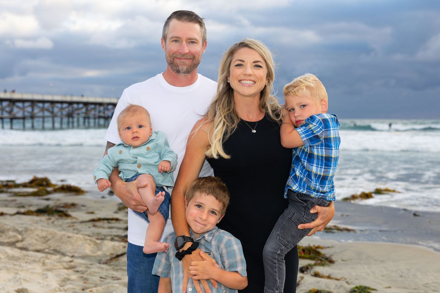 San Diego family portrait photography of a family of five posing on a beach with a wooden pier and ocean waves.