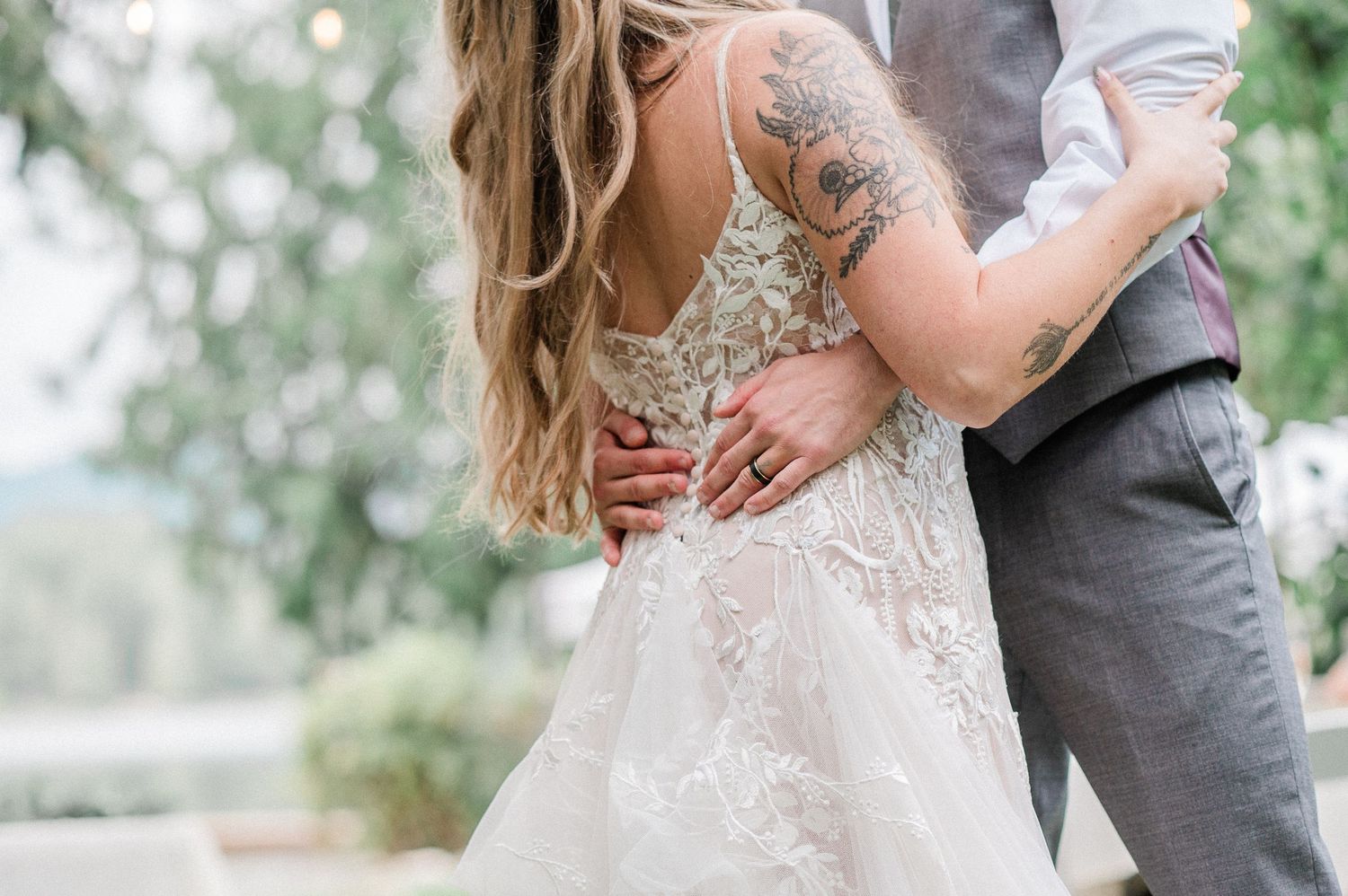 Bride in lace backless wedding gown embraced by groom in gray suit, showcasing floral arm tattoo outdoors.