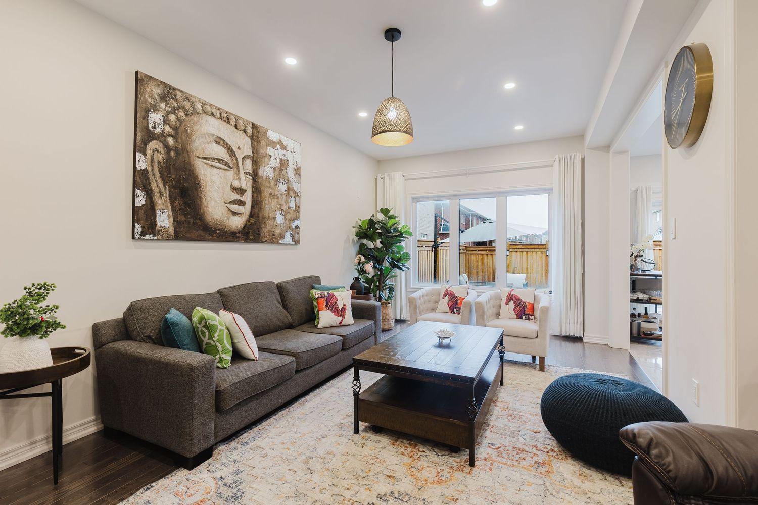 Modern living room with gray sofa, Buddha wall art, pendant light, and large window overlooking outdoor space.