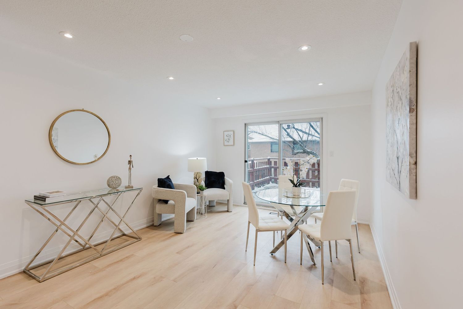 Bright modern living and dining area with light wood floors, glass dining table, white chairs, and large window.