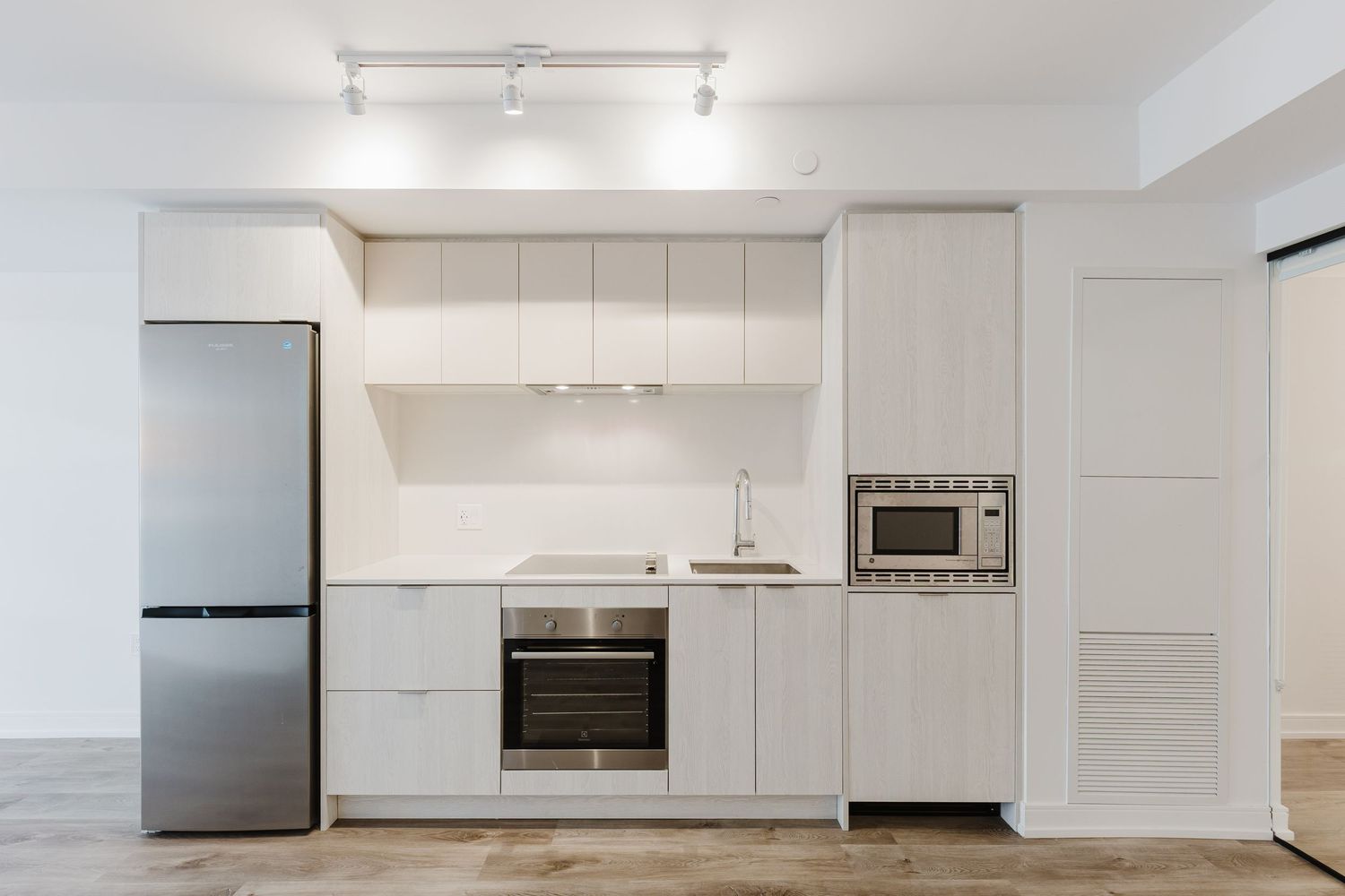 Modern white kitchen with stainless steel fridge, built-in oven, microwave, and sleek cabinetry on light hardwood floors.