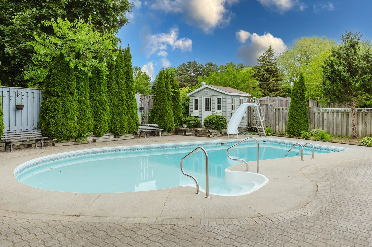 Backyard oasis featuring a large oval swimming pool with metal handrails, white slide, pool house, and lush green trees.