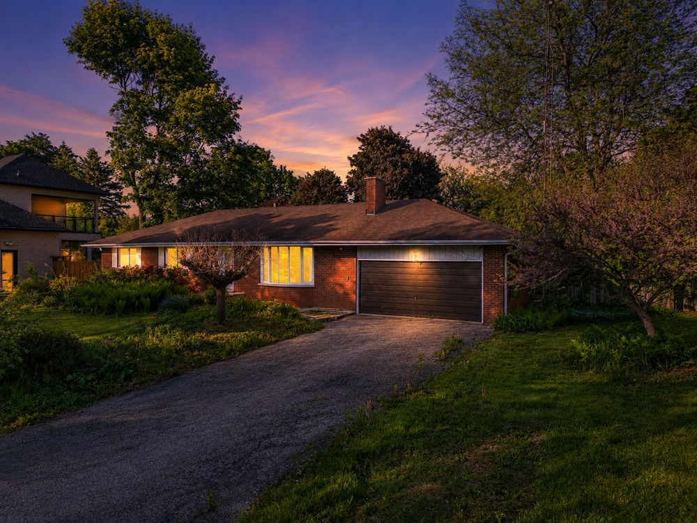Brick ranch home with attached garage at twilight, surrounded by lush green trees under a colorful sunset sky.