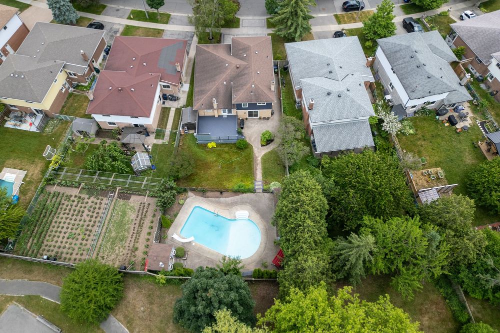 Aerial view of suburban backyard featuring a blue swimming pool, green lawn, garden beds, and surrounding neighborhood homes.