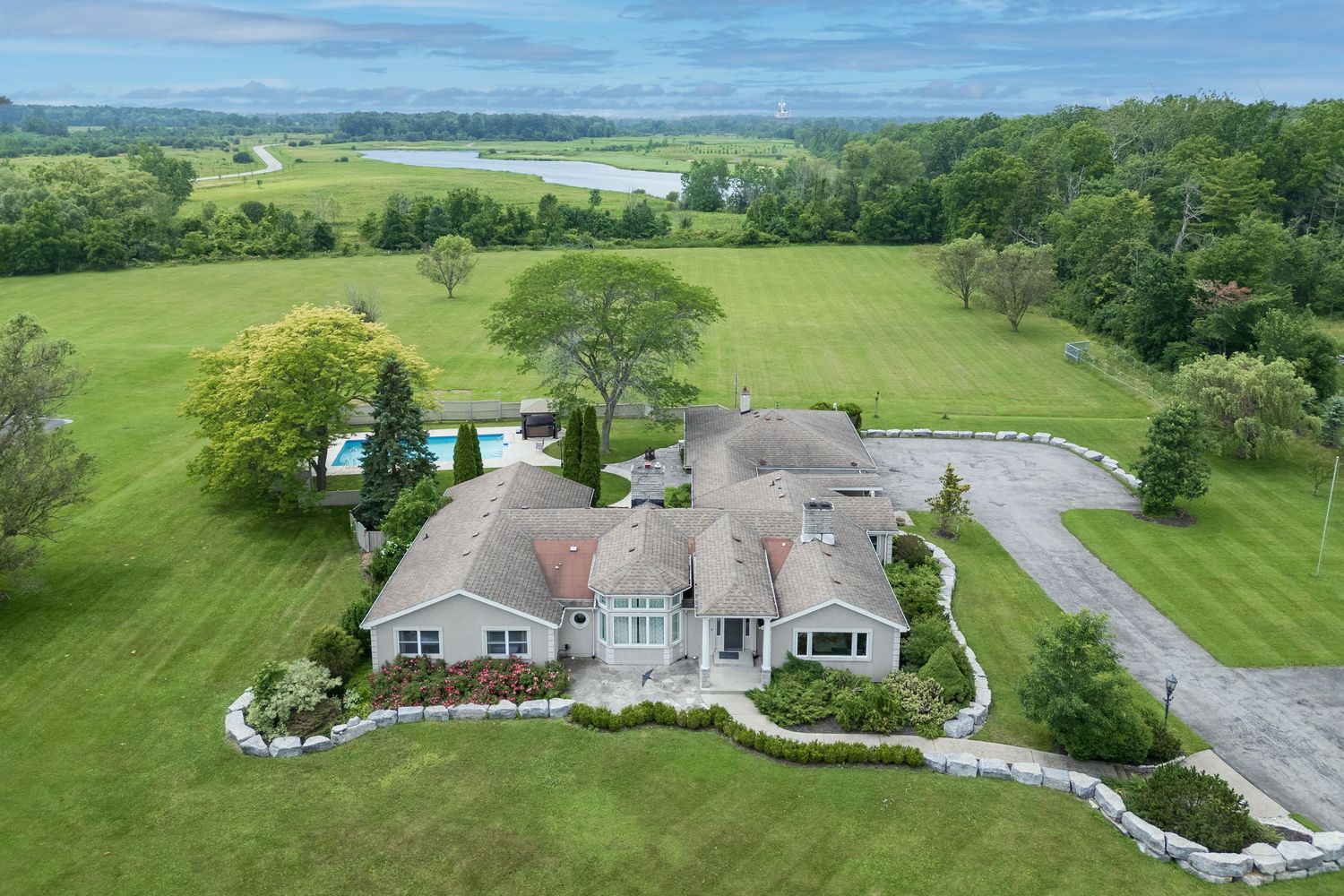 Aerial view of a sprawling ranch estate with lush green lawns, pool, circular driveway, and scenic water views beyond.