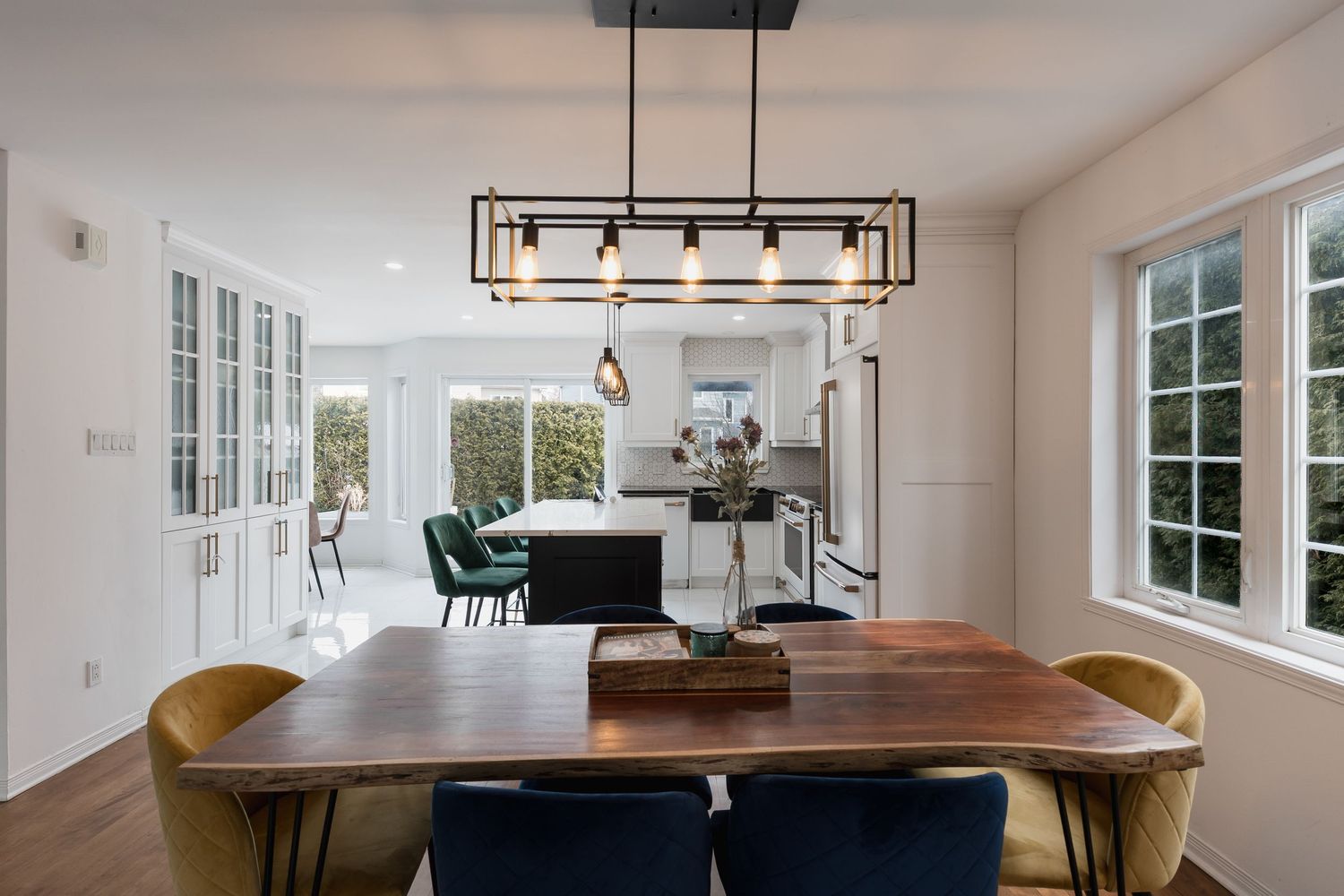 Modern dining room with wooden table, velvet chairs, and pendant light opening to a bright white kitchen.