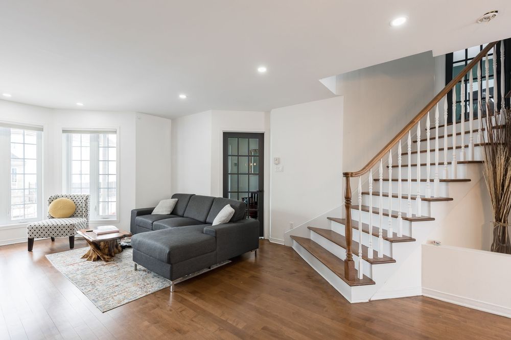 Bright living room with gray sectional sofa, hardwood floors, white staircase, and large windows in a modern home.