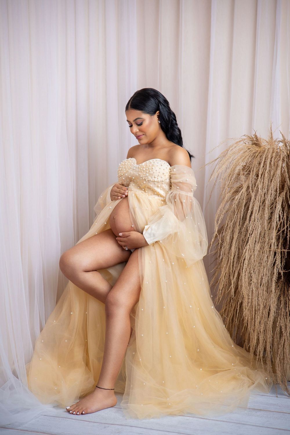 Pregnant woman in elegant gold tulle maternity gown posing in studio with neutral backdrop and dried pampas grass.