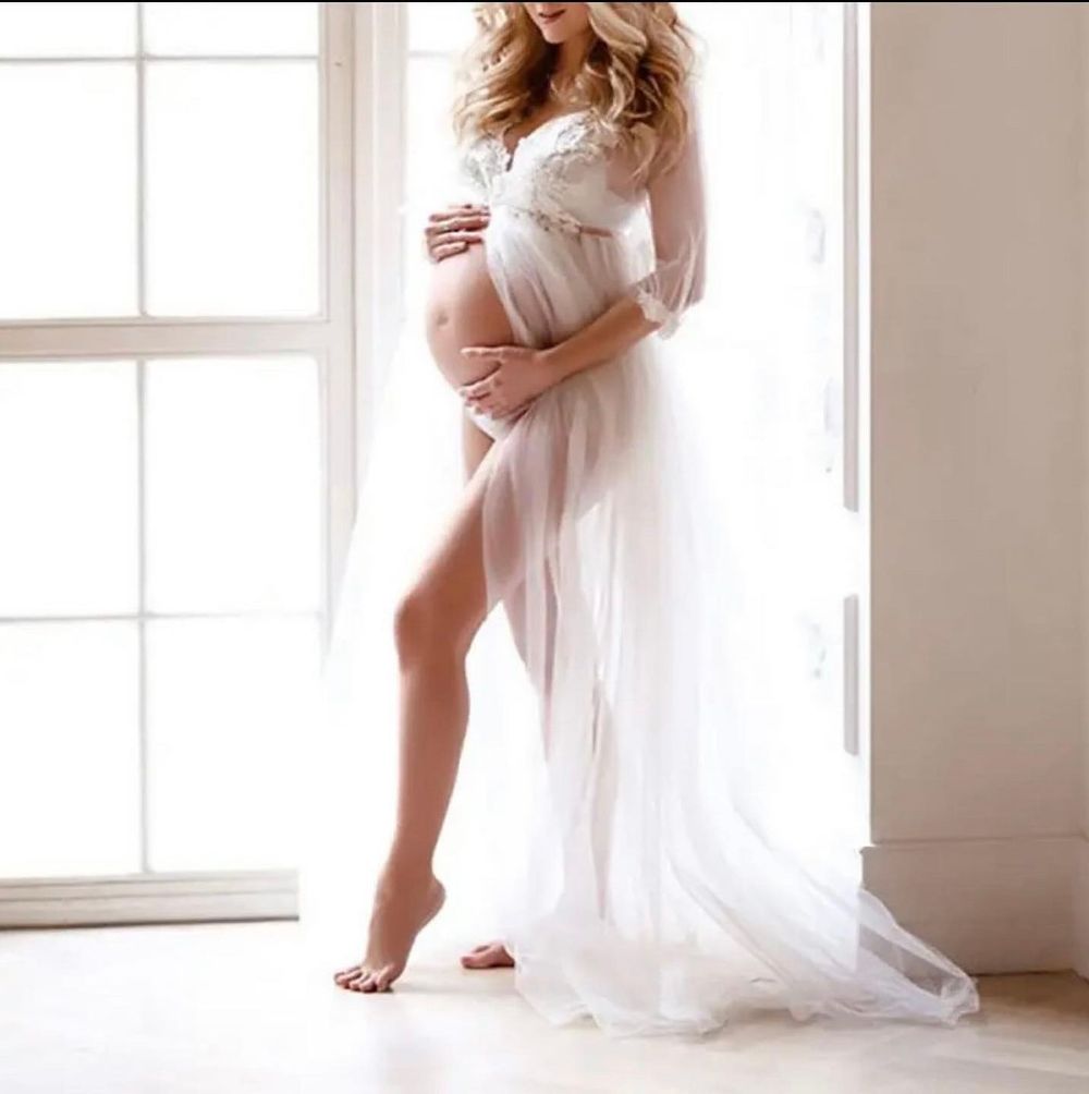 Pregnant woman in elegant white lace and sheer maternity gown posing gracefully by a bright window.