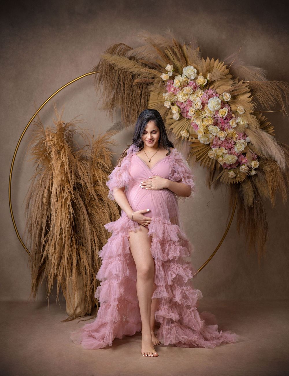Pregnant woman in pink ruffled maternity gown posing in front of floral hoop with pampas grass backdrop.