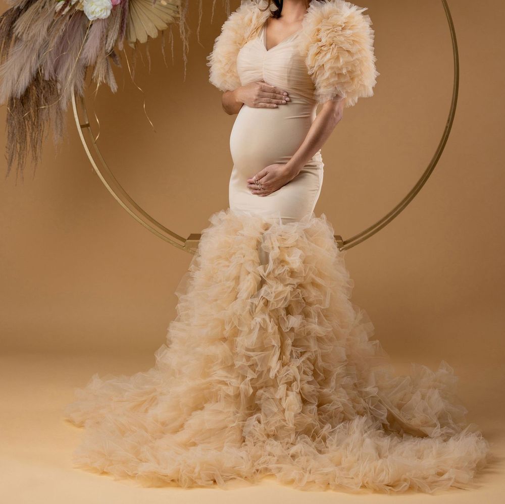 Pregnant woman in champagne mermaid maternity dress with ruffled tulle skirt posing before circular gold arch.