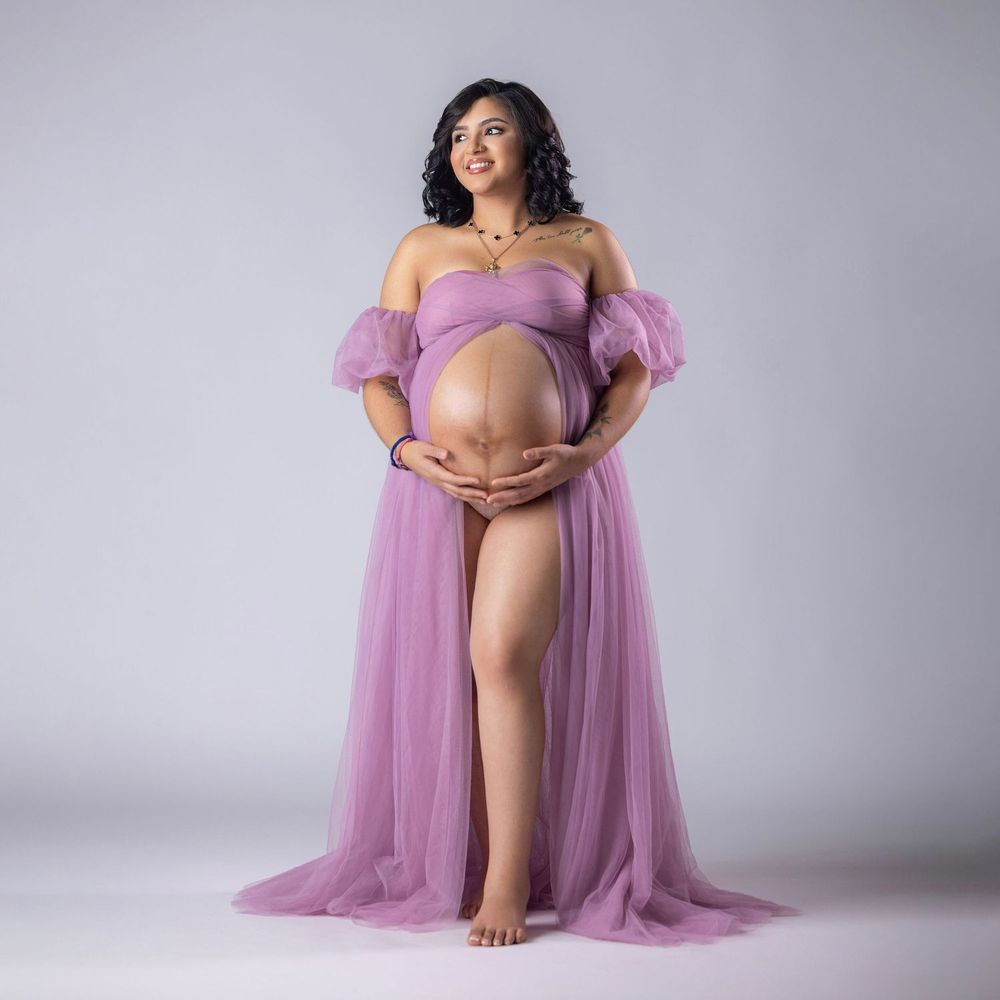 Smiling pregnant woman in off-shoulder purple tulle maternity gown with thigh-high slit in white studio.