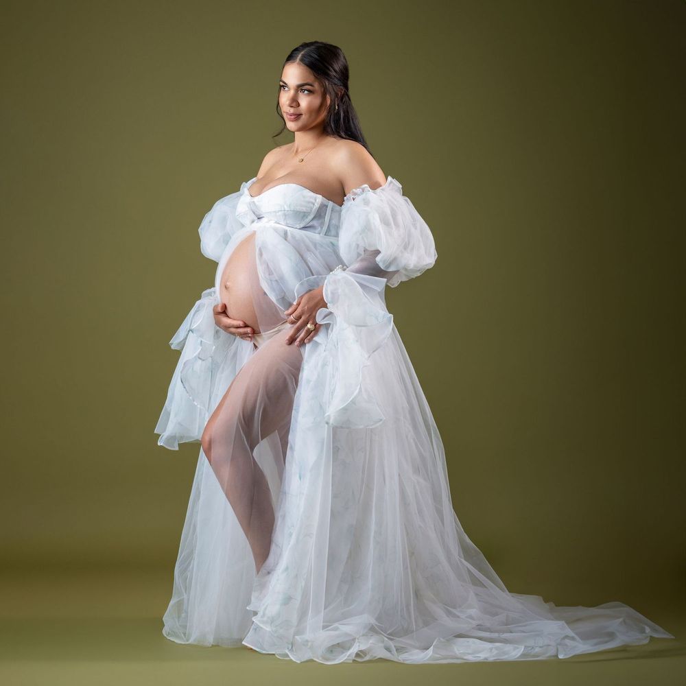 Pregnant woman in white off-shoulder tulle maternity gown posing elegantly against olive green background.