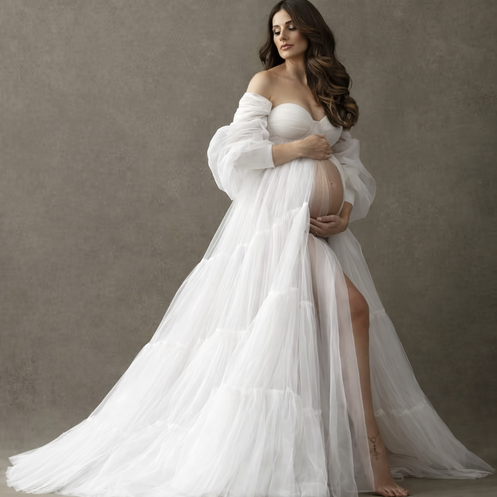 Pregnant woman in elegant white off-shoulder tulle maternity gown with puff sleeves and side slit for photoshoot.