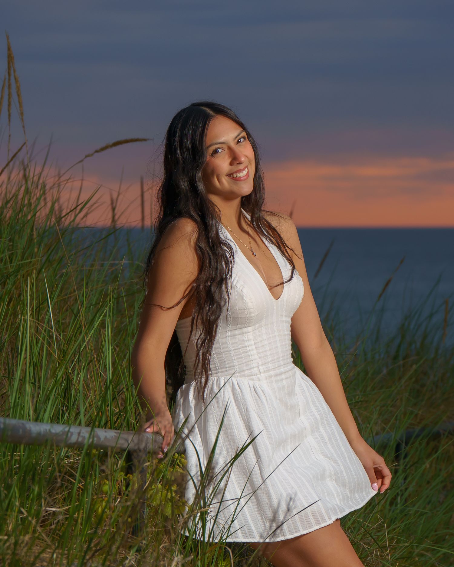 Young woman in white dress smiling at sunset beach, surrounded by tall grass with colorful sky backdrop.