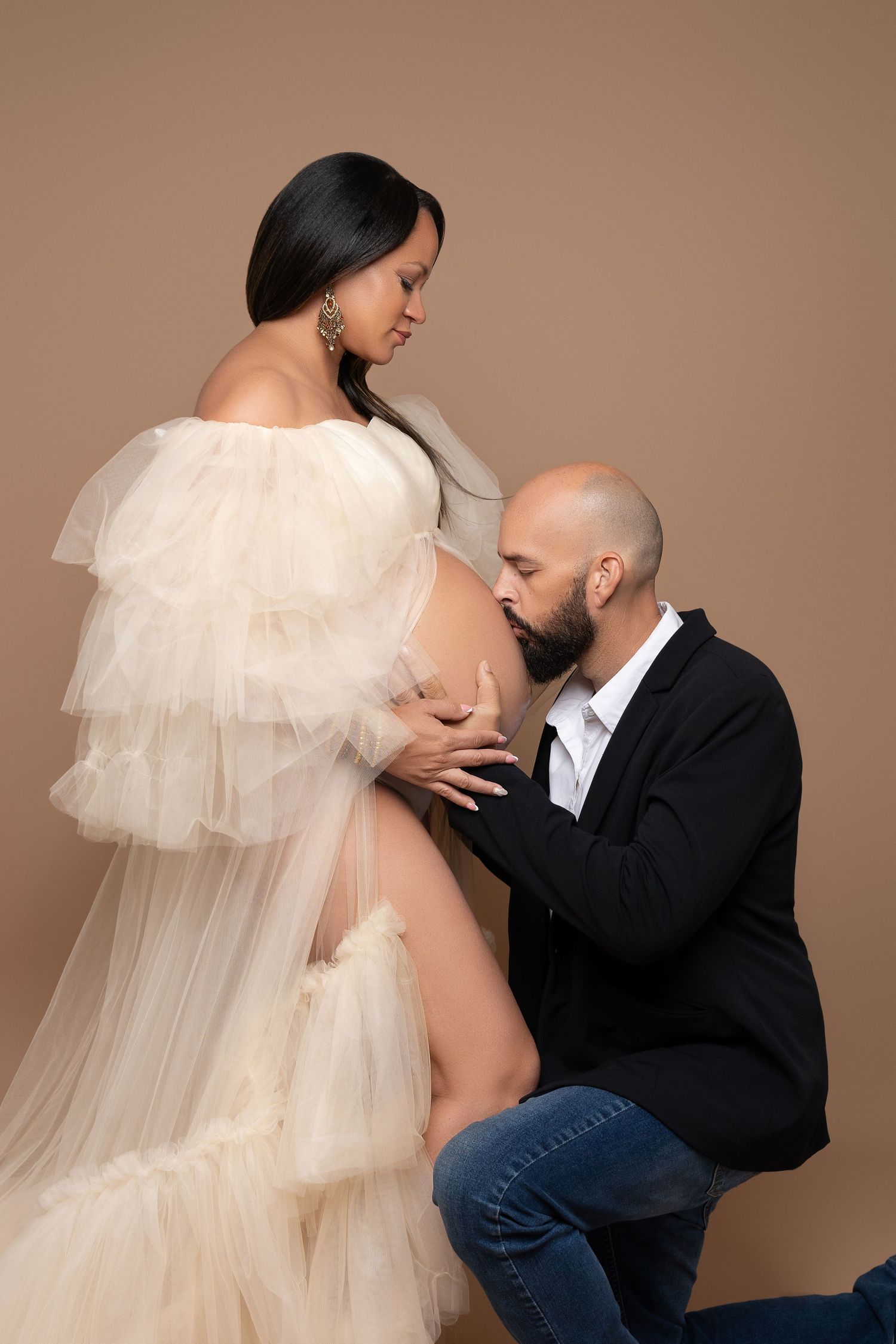 A pregnant woman in a white tulle gown poses with her partner kneeling beside her, kissing her baby bump in a studio.