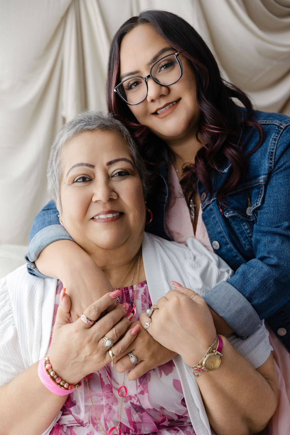 Two women embrace warmly, younger woman in denim jacket hugs older gray-haired woman wearing pink floral dress and bracelets.