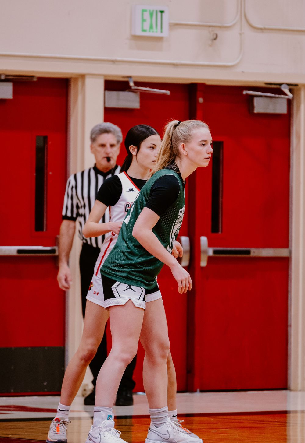 Players in defensive stance during basketball game with referee in background near exit door.