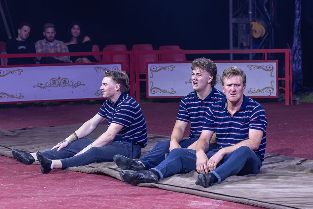Three performers in matching striped shirts sit on stage in synchronized pose.