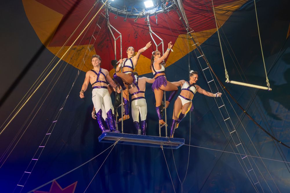 Circus performers execute a synchronized aerial routine on a suspended platform under colorful lighting.