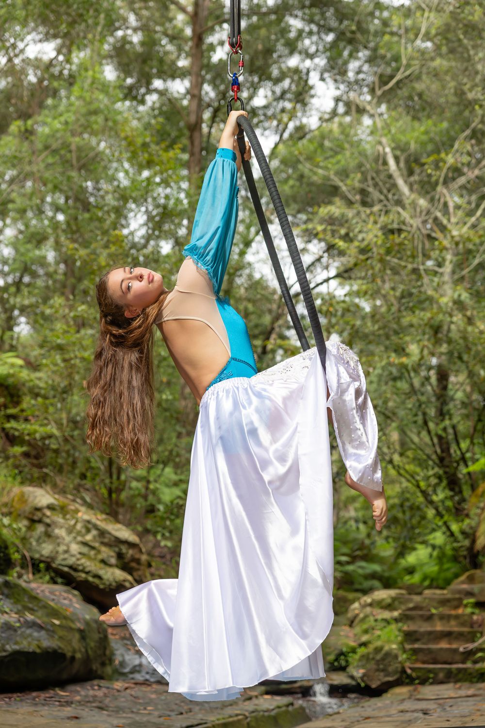 An aerial dancer in turquoise and white performs a graceful backbend pose while suspended from rigging in a forest setting.