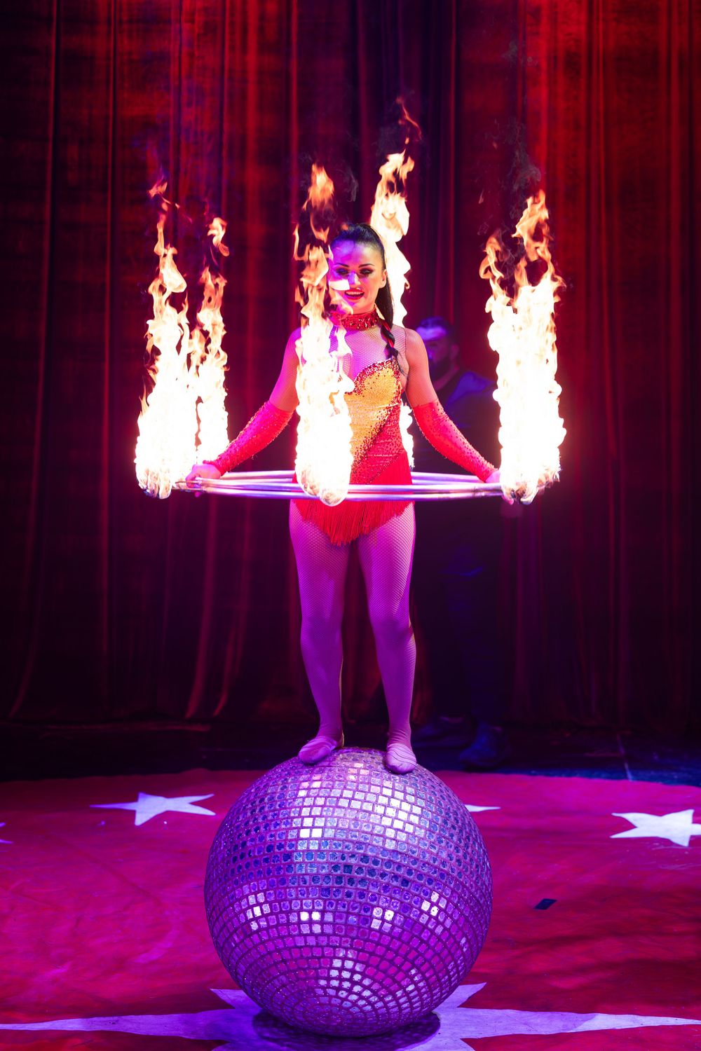 Circus performer balances on a glittering disco ball while holding a flaming hoop in a dramatic stage performance.