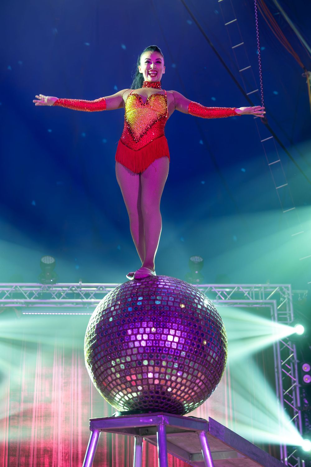 Circus performer balances on a large glittering disco ball under dramatic blue lighting on stage.