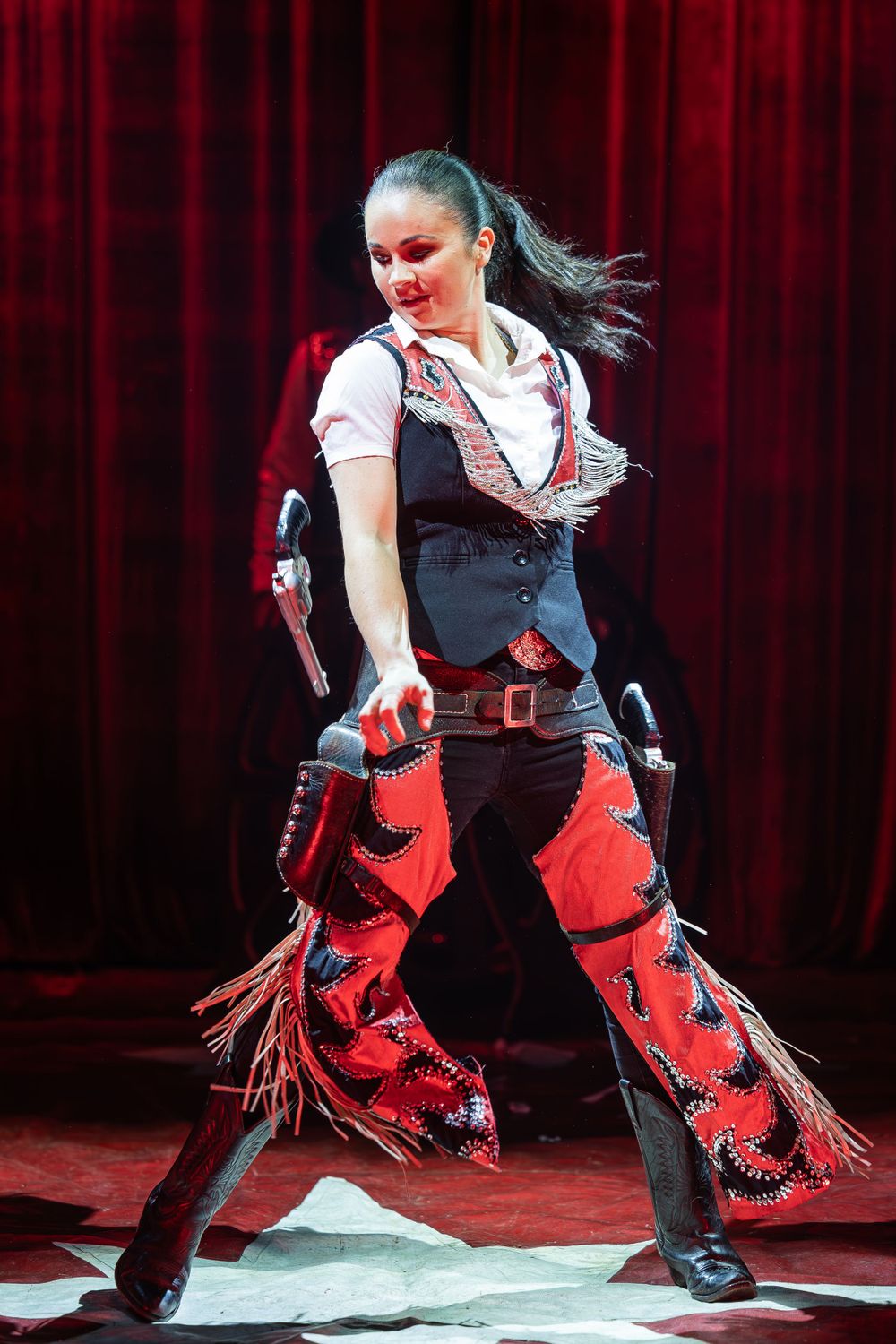 Performer in western-style costume with fringe chaps dances energetically on stage.