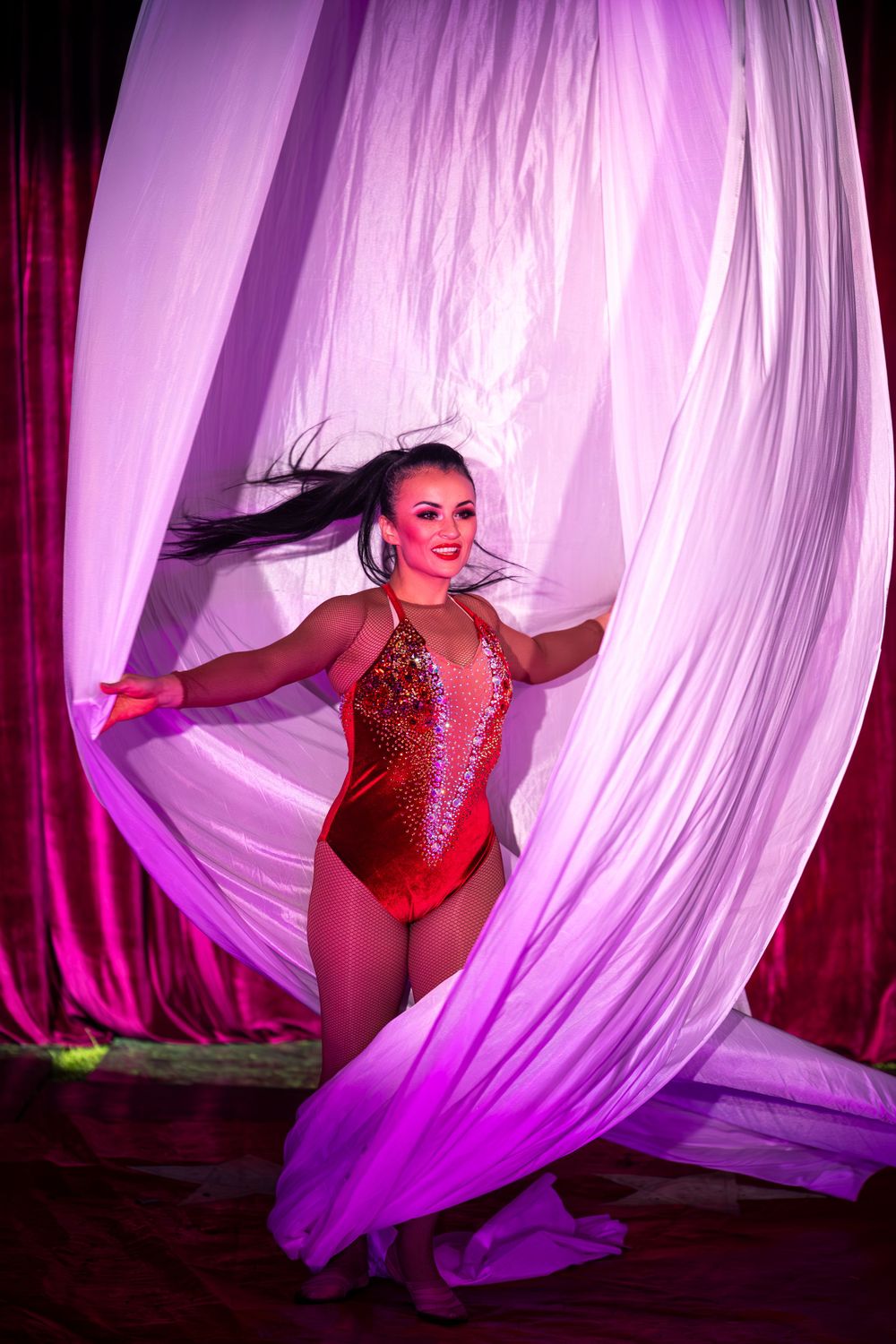 Performer in red sequined leotard poses gracefully between flowing pink silk curtains.