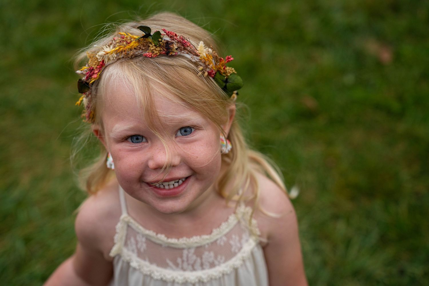 Adorable smiling blonde toddler girl wearing floral crown in white lace dress outdoors.