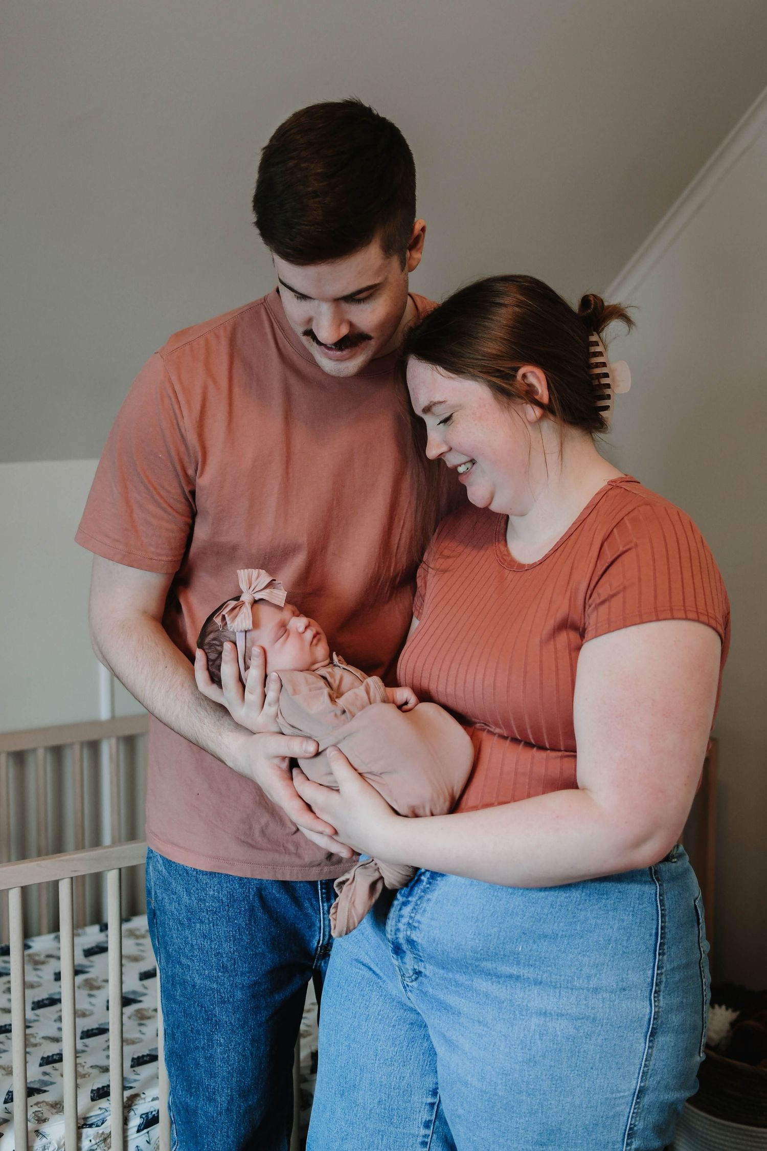 Parents in matching coral outfits share tender moment with newborn by crib.
