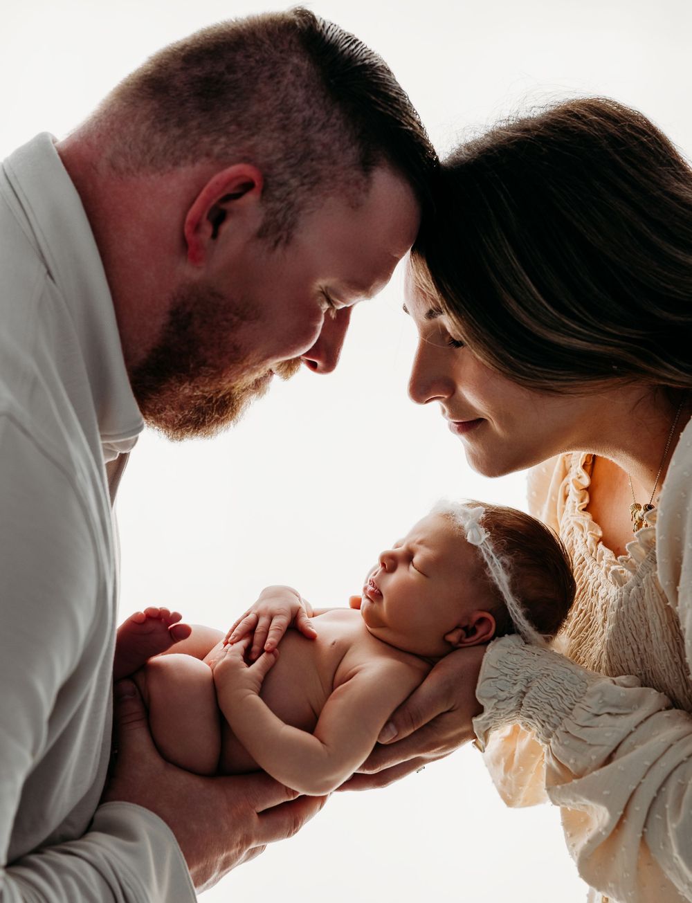 Parents lean in close and gaze lovingly at their newborn baby lying between them against white backdrop.