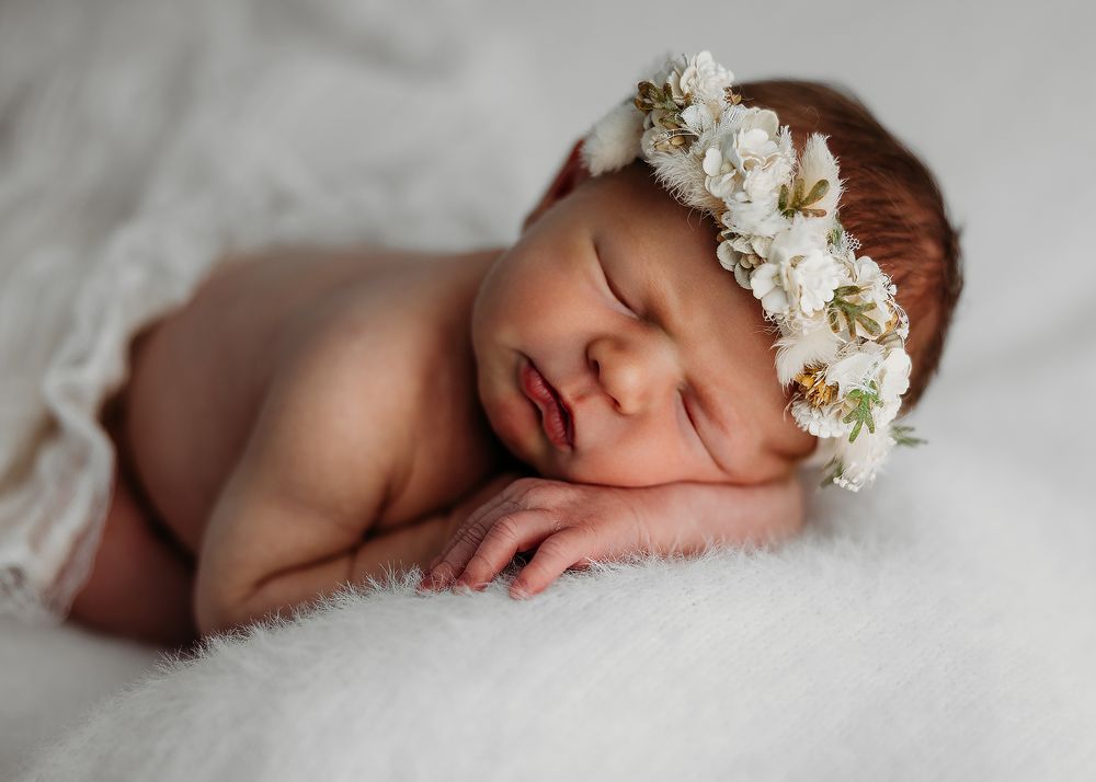 A sleeping newborn baby wearing a white floral crown rests peacefully on soft grey fabric in a delicate portrait series.
