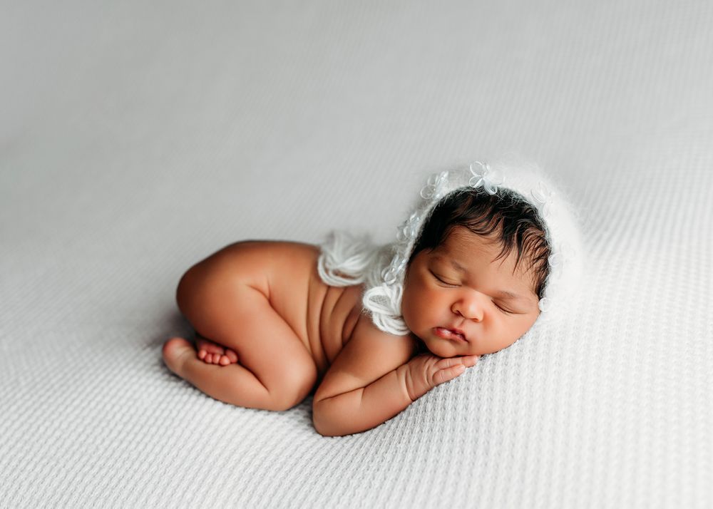 Sleeping newborn baby wearing white bonnet rests peacefully on textured white blanket in curled position.