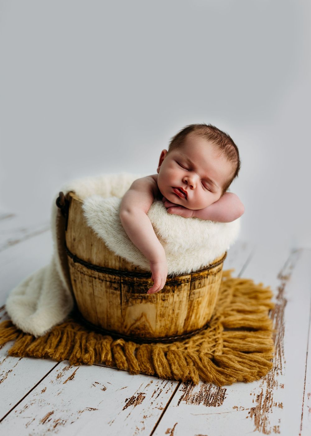 Sleeping newborn rests peacefully in rustic wooden barrel on white fluffy blanket and textured brown mat.