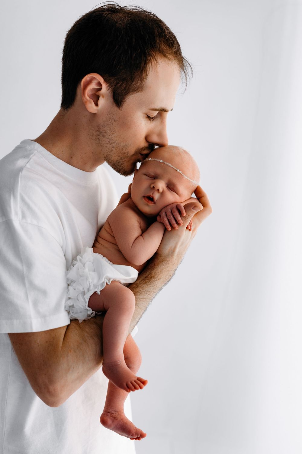 A tender moment showing an adult in a white t-shirt gently cradling and kissing a sleeping newborn baby.
in Fairfax, VA -Nova photographer